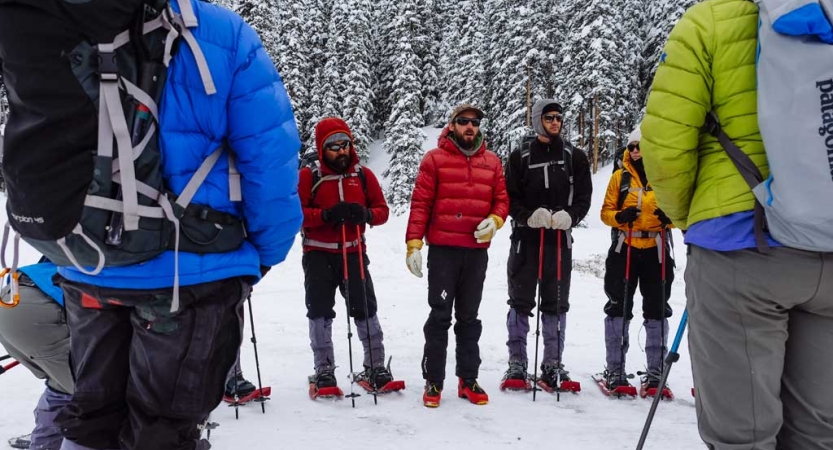 a group of veterans wearing snow gear and snowshoes stand in a circle while an instructor speaks